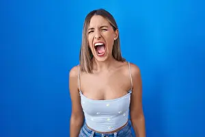 Evil Prints: Young woman standing over blue background angry and mad screaming frustrated and furious