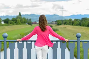 Contemplative Prints: Young woman standing looking out over a rural river in lush green countryside leaning on the blue metal railings of