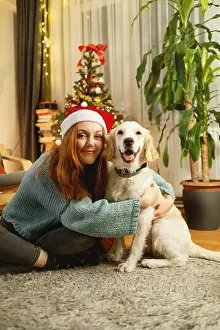 Cuddling Prints: A young woman wearing a Santa hat lovingly hugs her golden retriever while sitting on the floor
