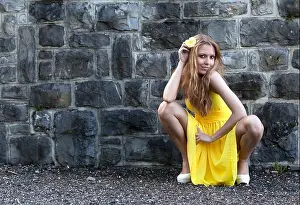 Poses Prints: Young woman in a yellow dress and a yellow flower posing in a crouching position in front of a stone wall